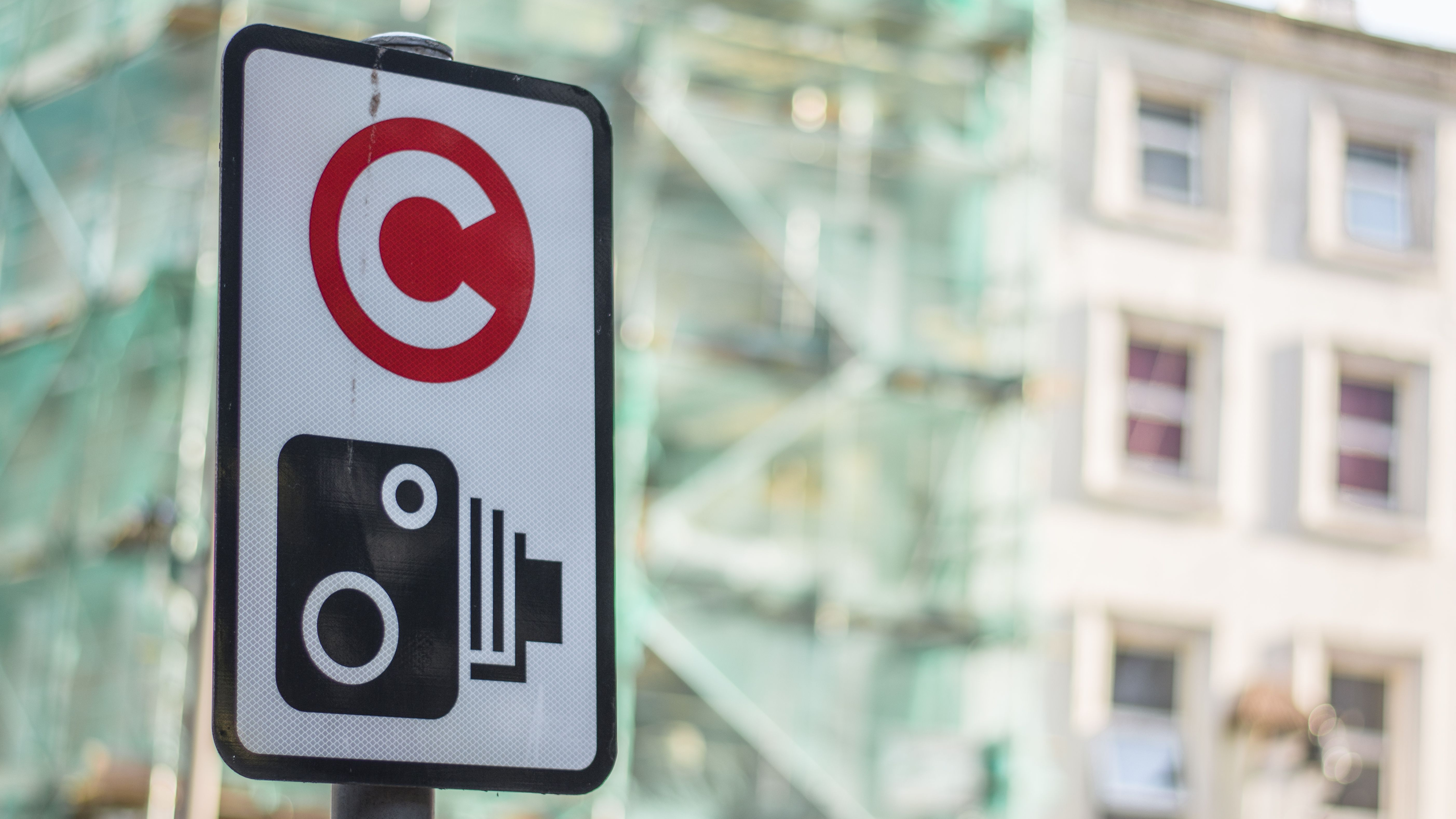 A close-up of a London Congestion Charge sign featuring a red 'C' symbol above a black camera icon. The sign is mounted on a pole, with a blurred background of a city street, buildings, and scaffolding