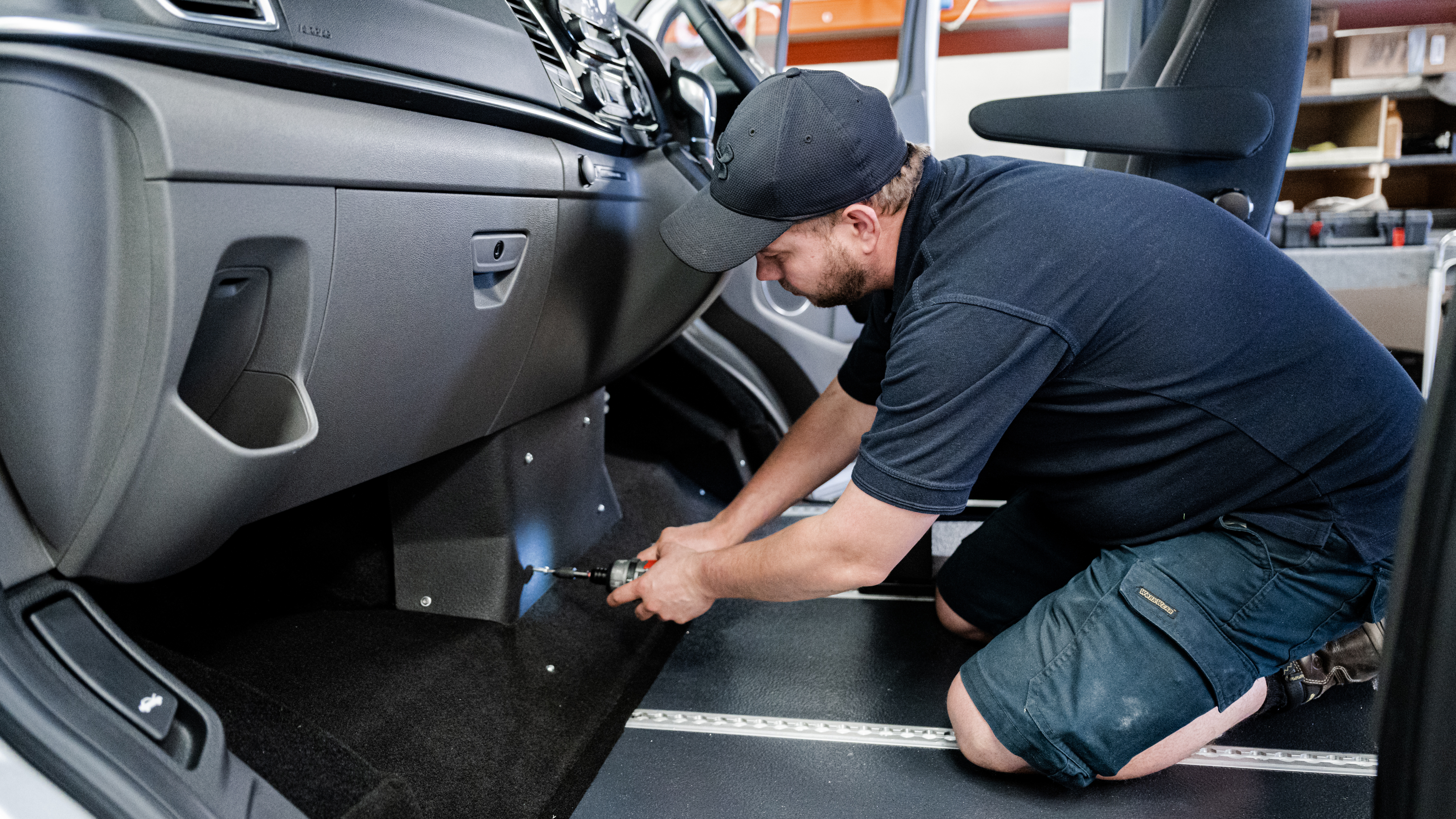 A mechanic is kneeling inside a vehicle, using a power tool to install or adjust a component beneath the dashboard on the passenger side. He is wearing a black cap, navy shirt, and work shorts, and appears focused on the task. The interior of the vehicle is clean and organized, with visible flooring modifications, possibly for accessibility or custom conversion purposes.