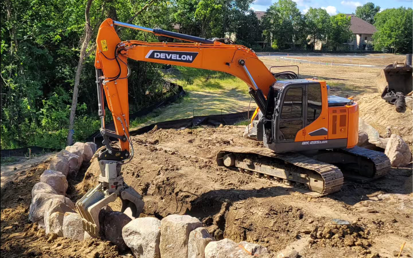 A DEVELON excavator positions boulders for a retaining wall with a grapple attachment.