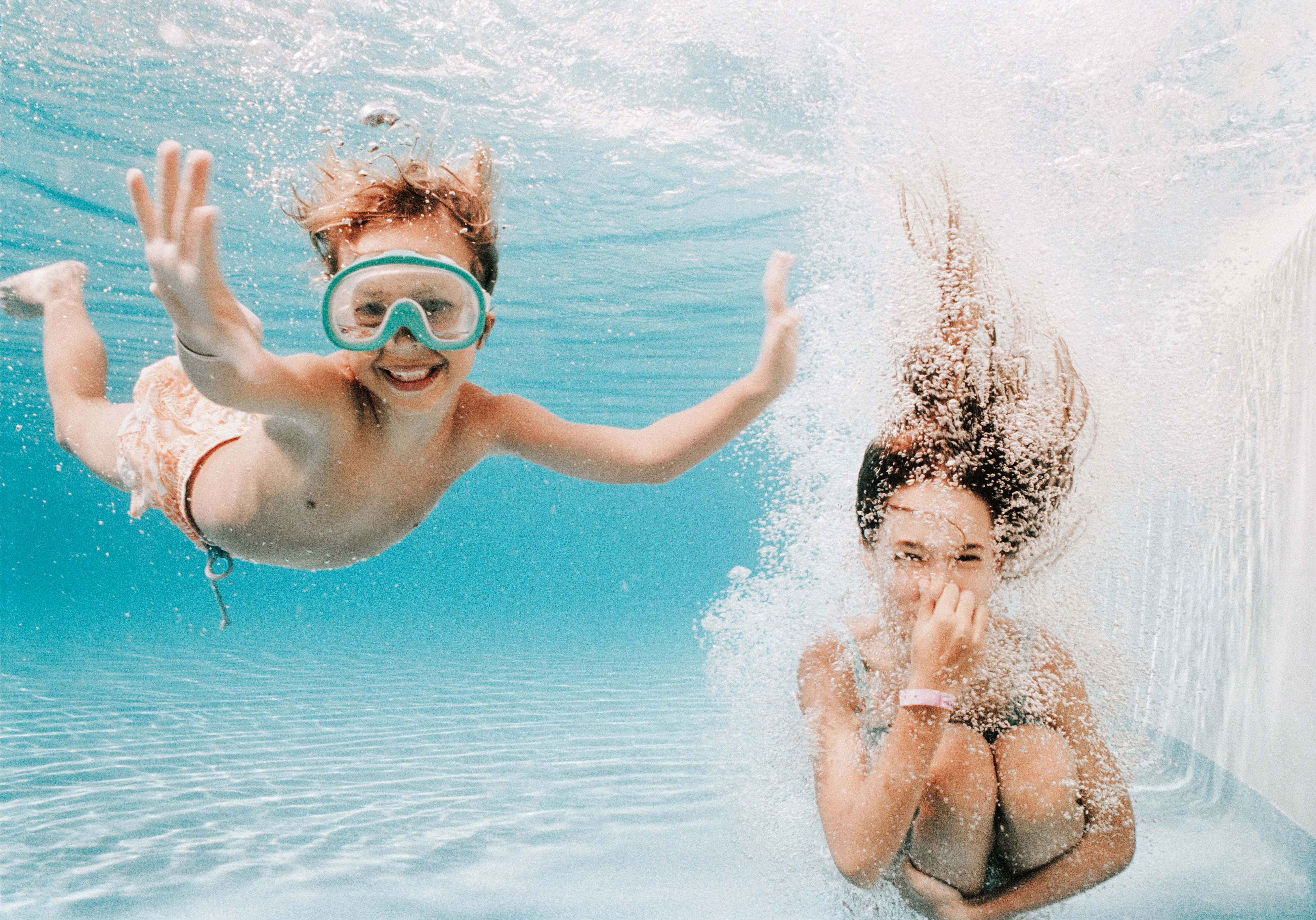 Two children underwater in a swimming pool, one wearing swim goggles. 