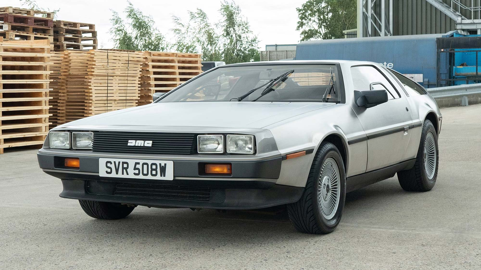 A classic silver DeLorean DMC-12 sports car with gullwing doors closed, parked near stacked wooden pallets in an industrial setting. UK registration plate reads SVR 508W.