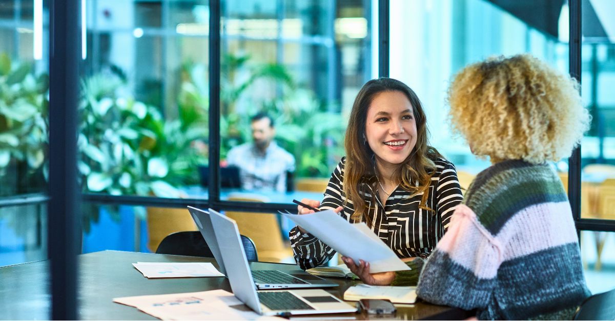 Two women in a meeting using a laptop and reviewing paperwork