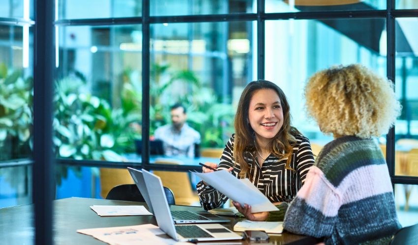 Two women in a meeting using a laptop and reviewing paperwork