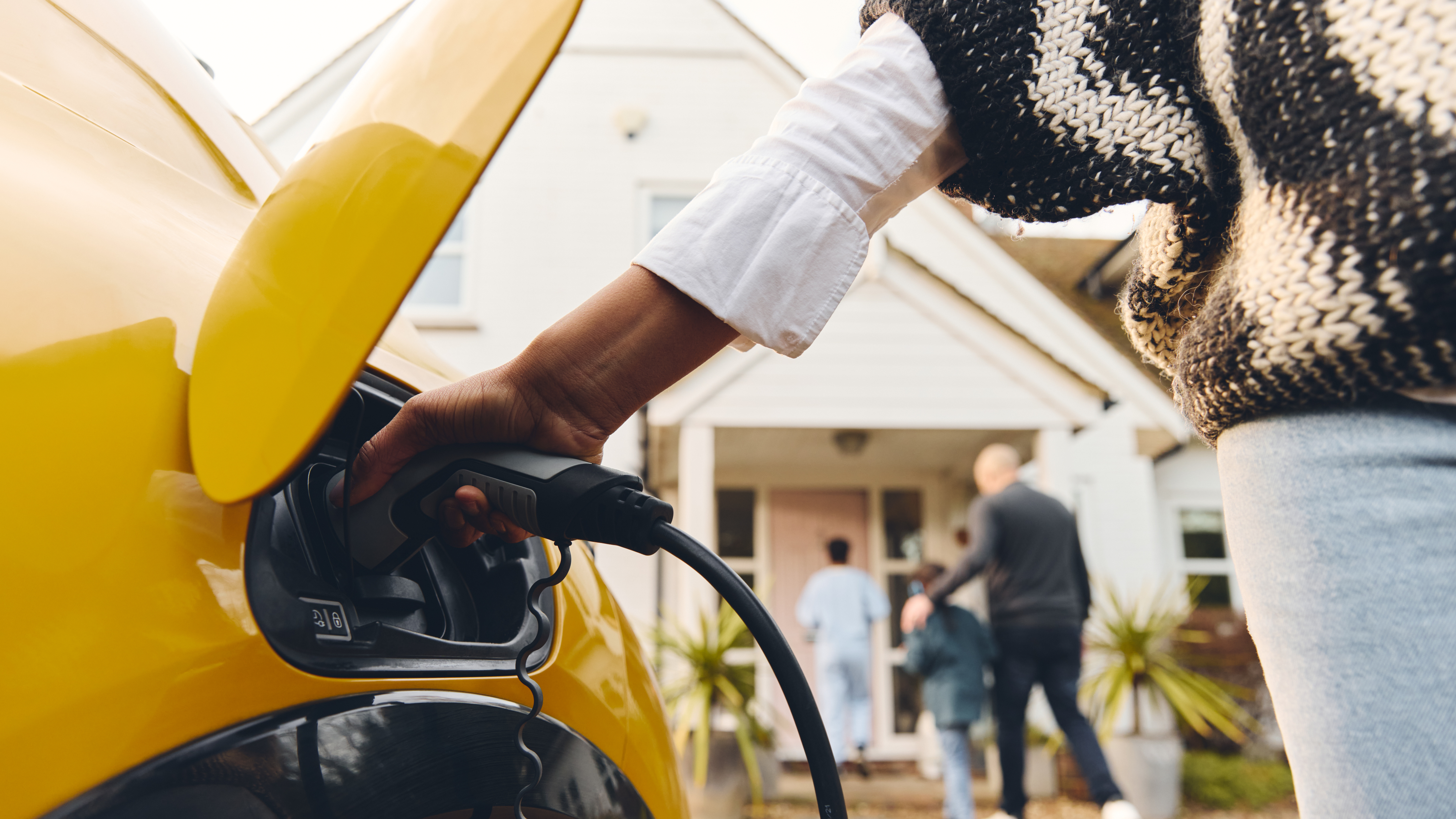 A close-up of a person plugging a charging cable into a yellow electric vehicle parked outside a house, with a family walking toward the front door in the background.