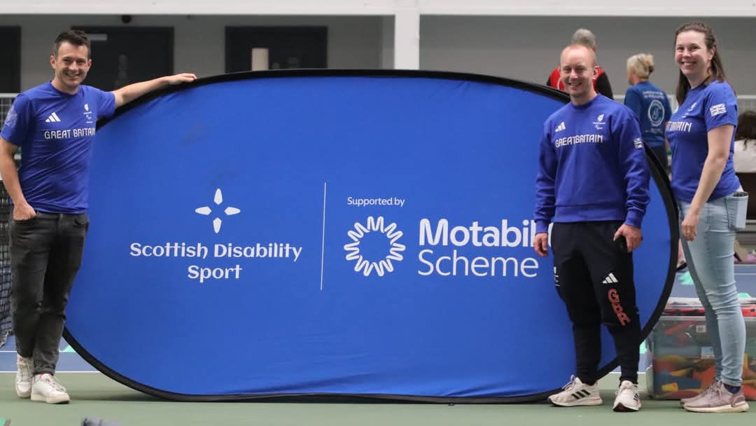 Three individuals in Team Great Britain attire stand beside a large blue banner displaying the logos of Scottish Disability Sport and the Motability Scheme at an indoor venue.