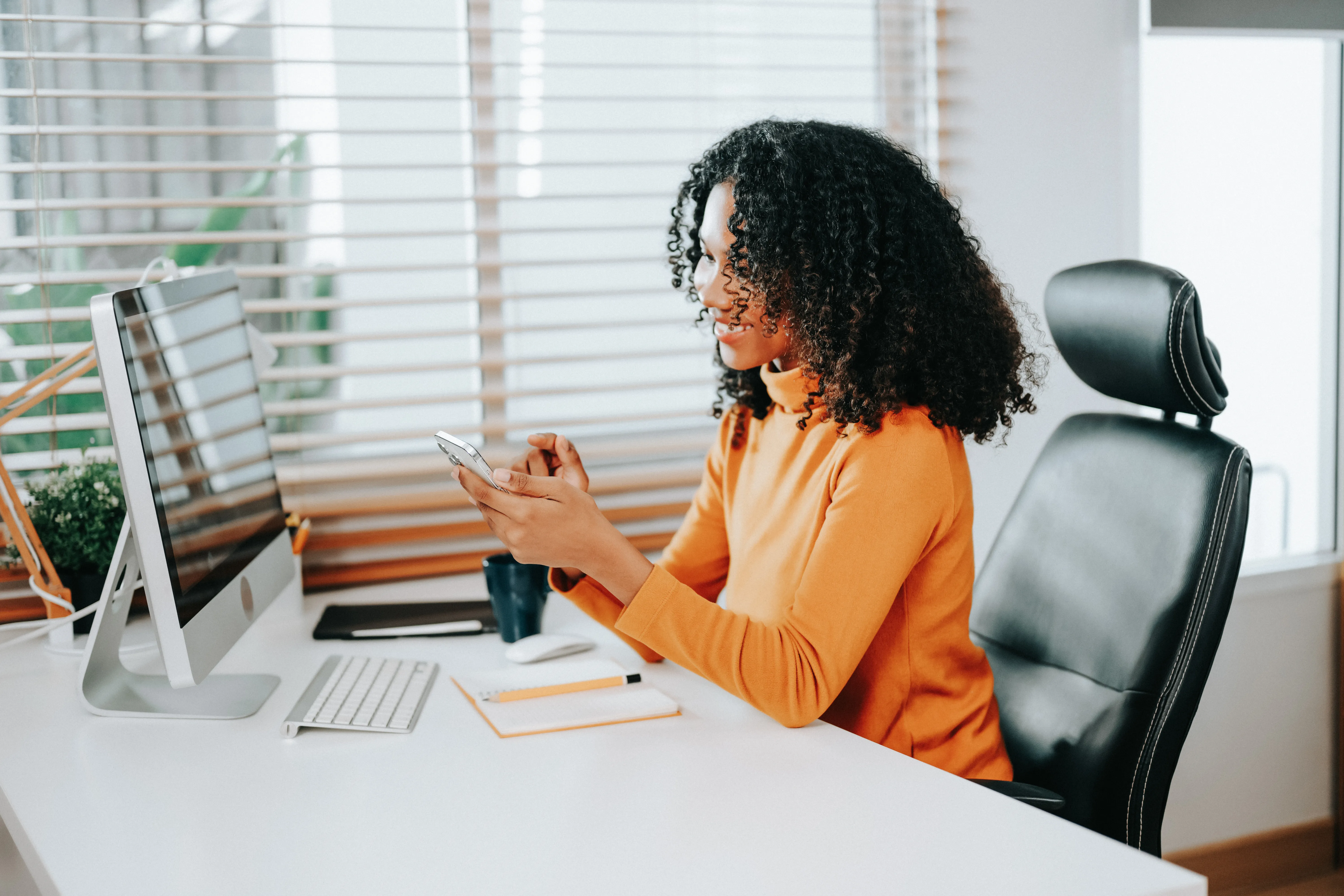 Professional African American woman sitting at desk in office looking at cell phone 
