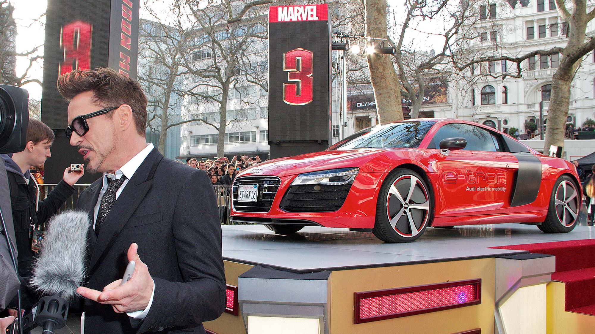 Robert Downey Jr. gives an interview on the red carpet at the Iron Man 3 premiere, with a red Audi R8 e-tron electric sports car displayed behind him and Marvel branding overhead.
