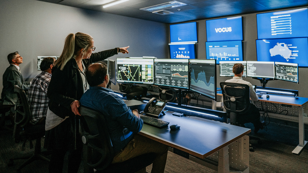 A Vocus security operations control room with multiple people working at computer stations. Several large monitors on the walls display various data visualisations, charts, and maps. One person is standing and pointing towards one of the screens, while others are seated and focused on their tasks. 