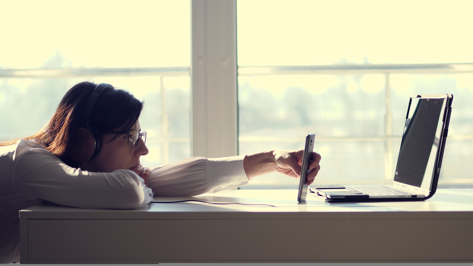 Lazy young woman, office worker wearing headphones, playing mobile games with smartphone during work hours. Bored employee. Useless and unproductive worker doing nothing and forget her job.