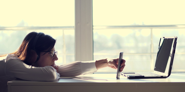 Lazy young woman, office worker wearing headphones, playing mobile games with smartphone during work hours. Bored employee. Useless and unproductive worker doing nothing and forget her job.