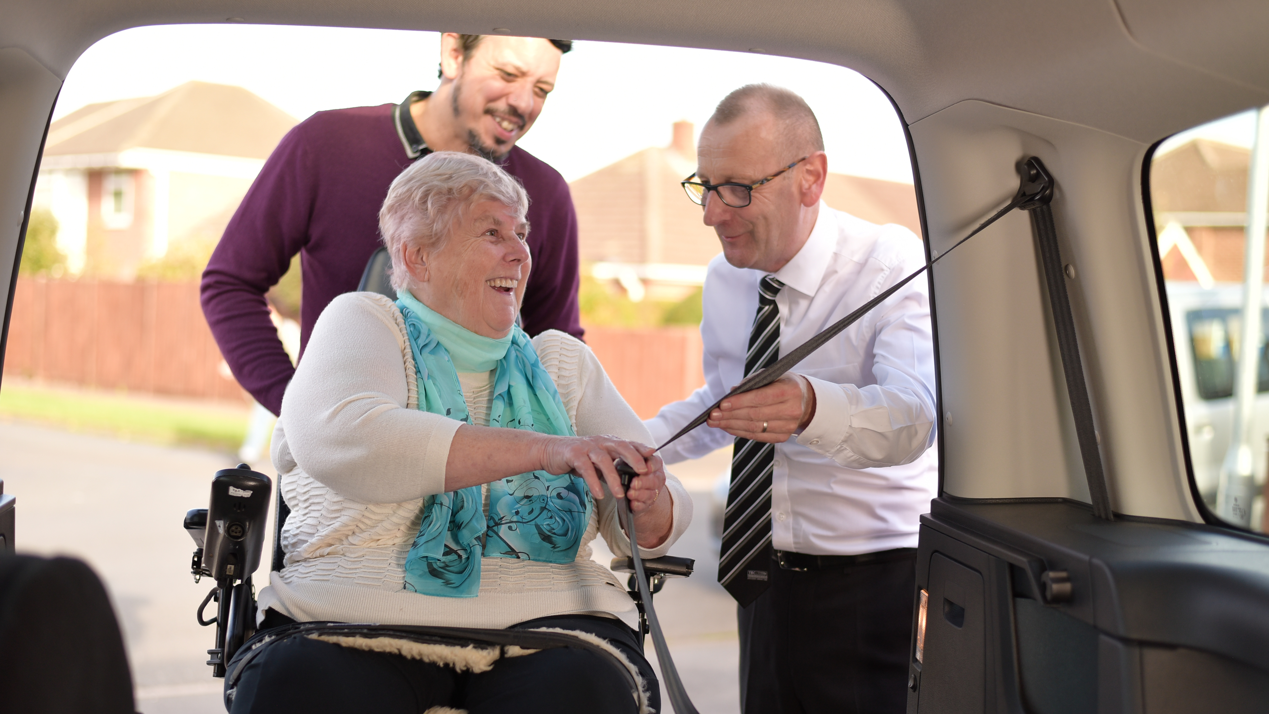 An older woman in a powered wheelchair smiles as two men assist her inside a vehicle, with one man demonstrating how to secure the seatbelt.