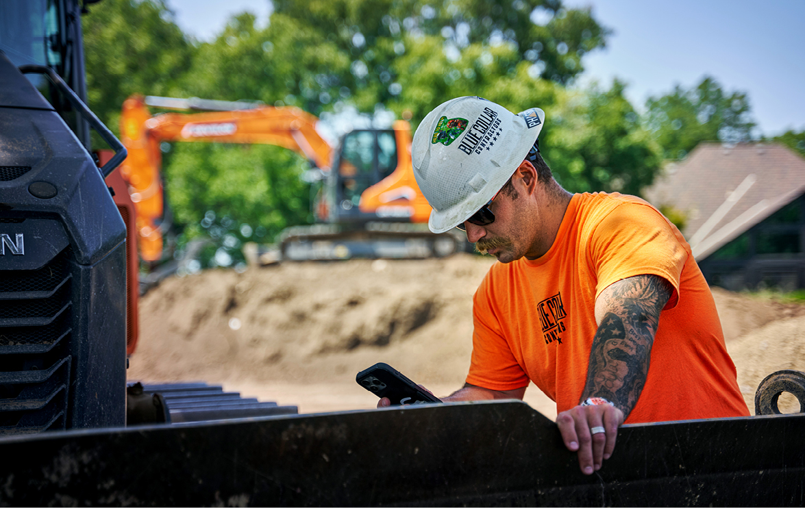 Trent Harris, director of operations at Blue Collar Contractors, looking at his smartphone with a DEVELON DX140LC-7 crawler excavator in the background on a job site. 
