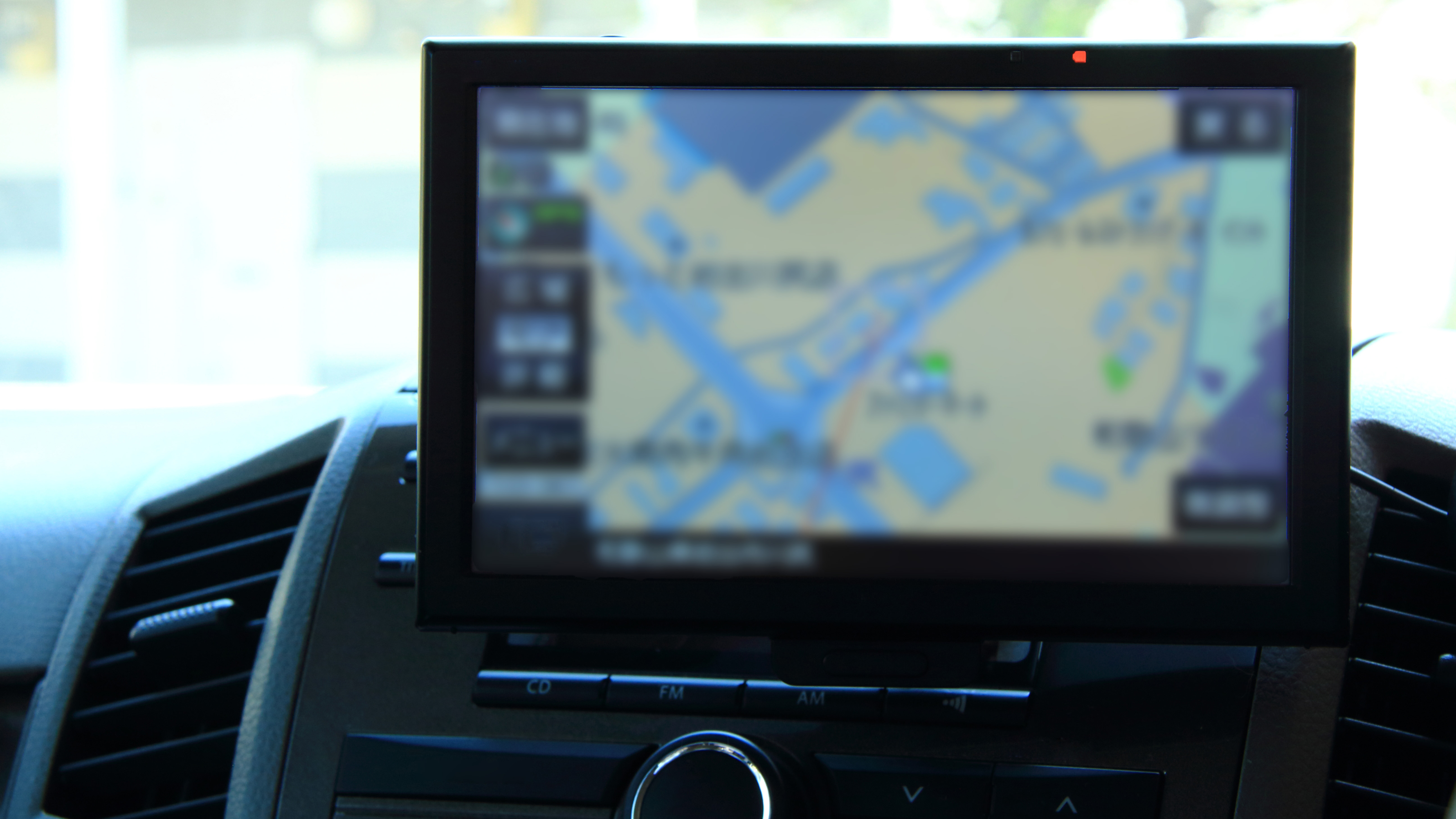 A close-up of a car's dashboard navigation system showing a digital map on the screen. The display is slightly blurred, but roads and locations are visible. The device is mounted in the centre console, surrounded by air vents and control buttons.