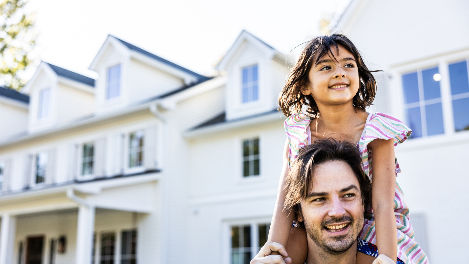 Young girl riding on her fathers shoulders in front of suburban home