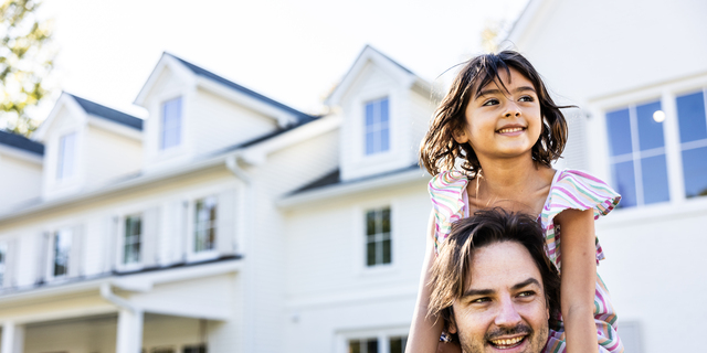 Young girl riding on her fathers shoulders in front of suburban home