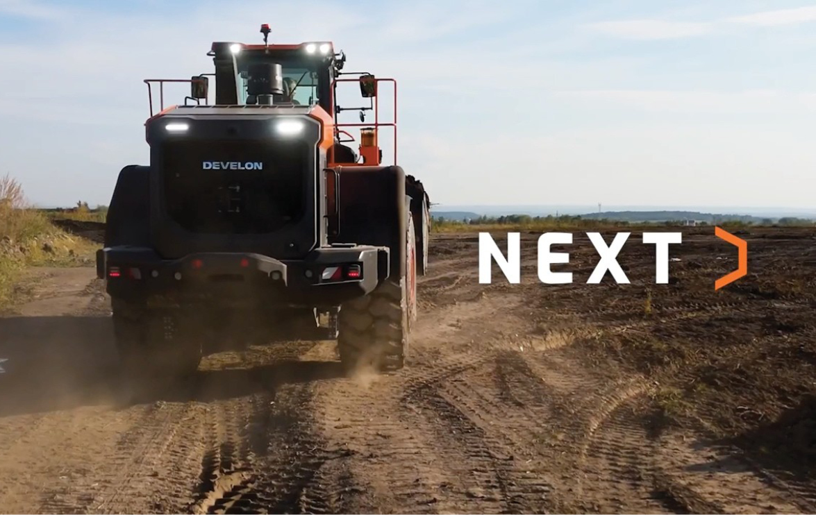 A DEVELON wheel loader driving away on a dirt jobsite with the NEXT logo to its right