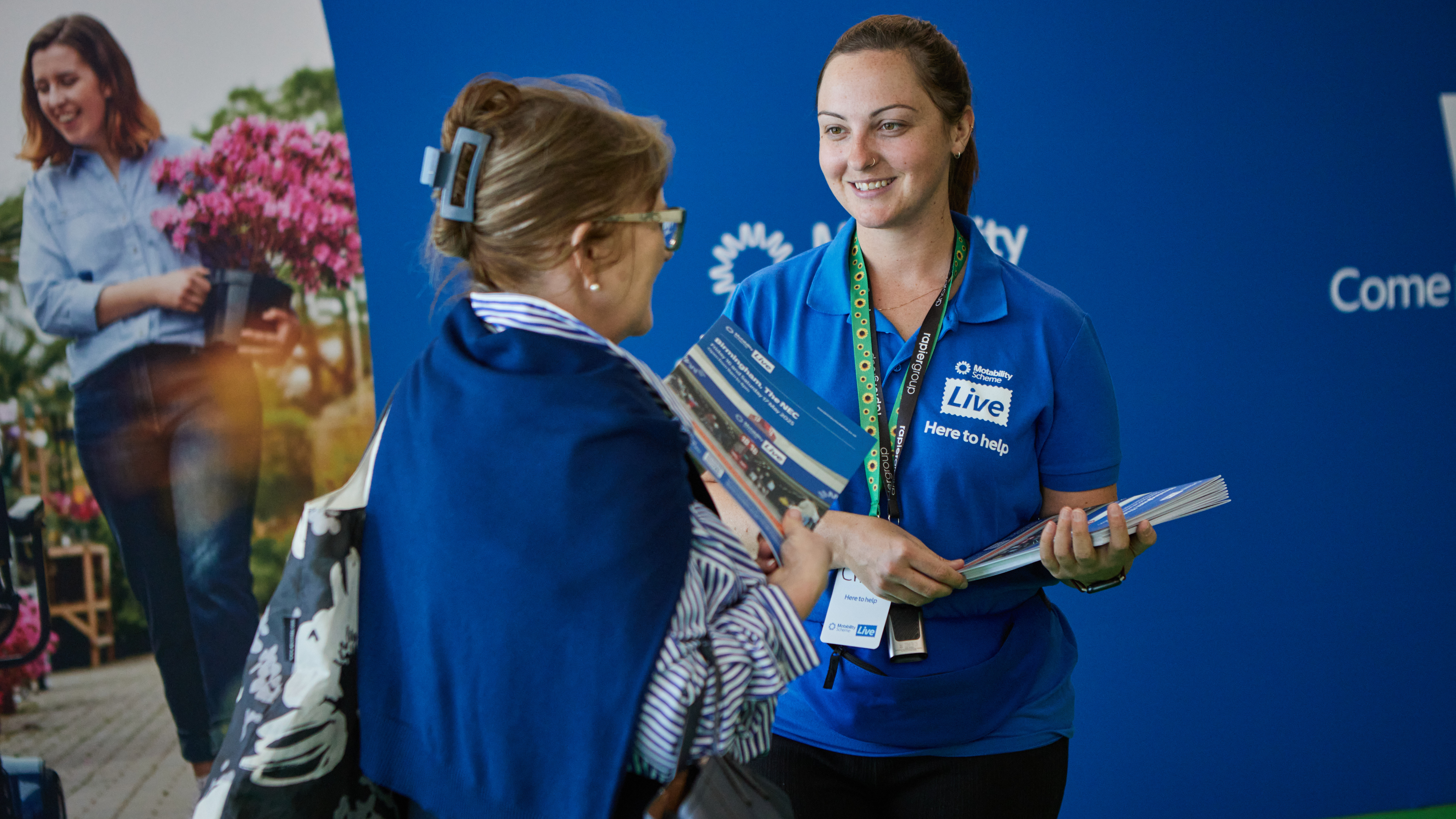 A Motability Scheme staff member in a blue “Here to help” shirt hands out brochures and smiles while assisting a visitor at an event.