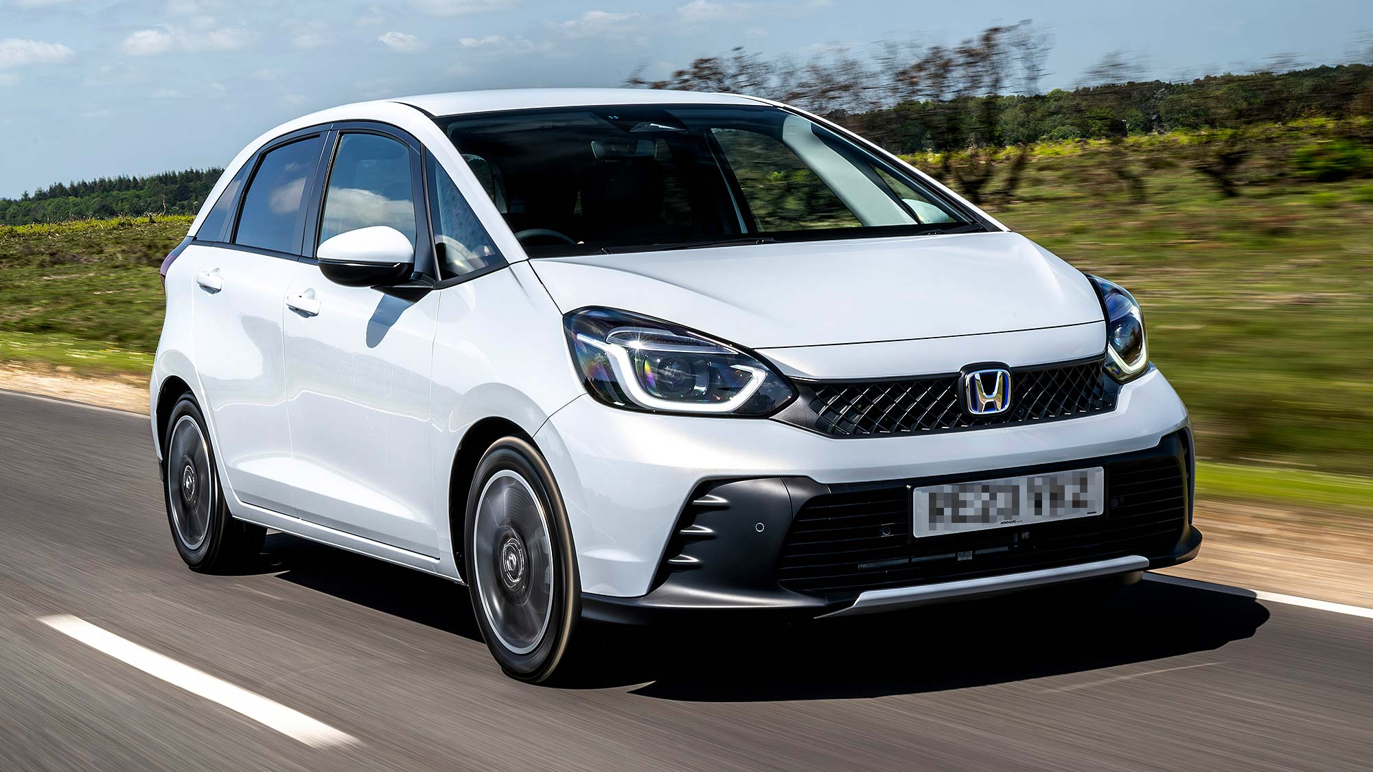 A white Honda Jazz hybrid hatchback driving on a countryside road, with a clean design and compact proportions.