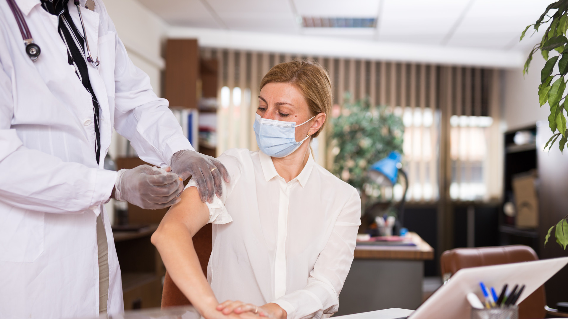 Doctor in coat and protective mask giving vaccine injection to woman manager
