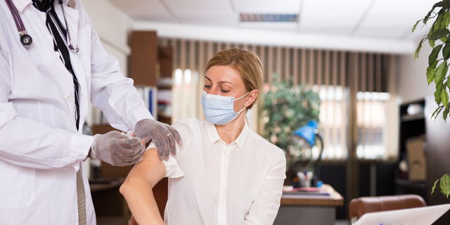 Doctor in coat and protective mask giving vaccine injection to woman manager