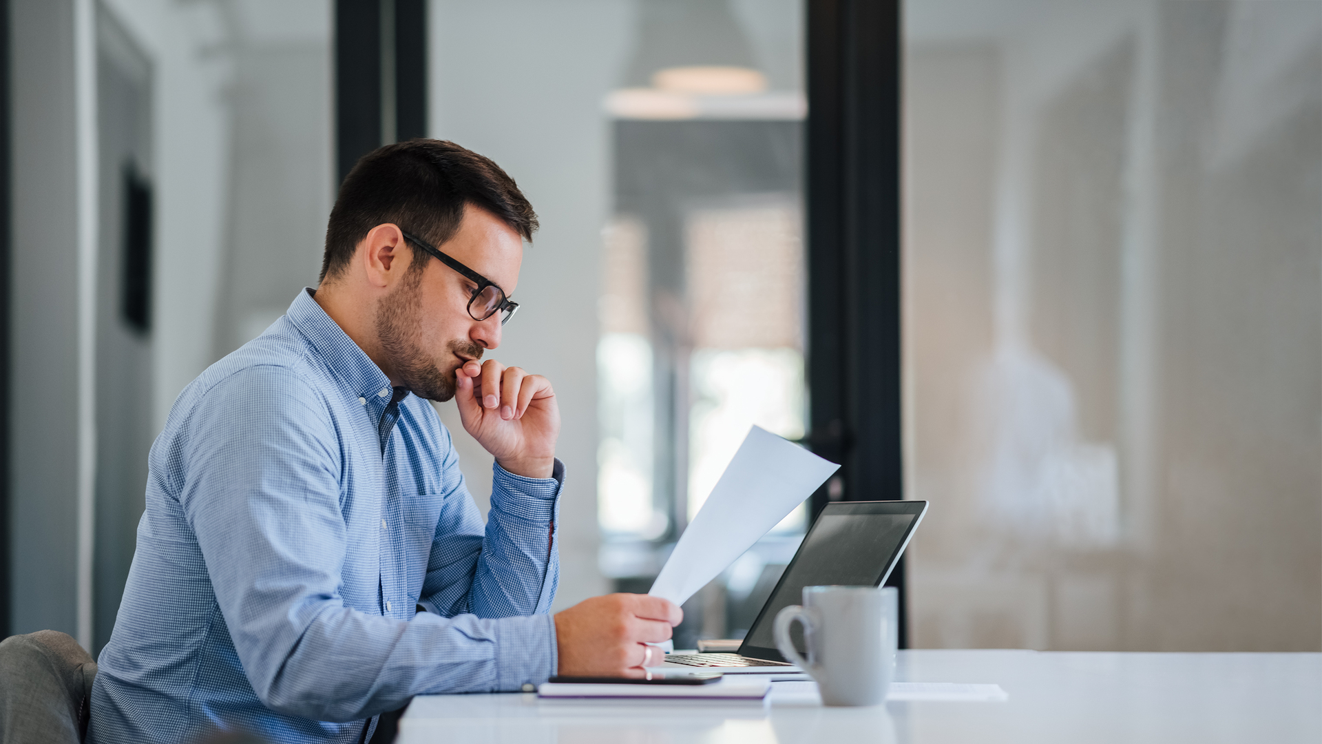 Serious pensive thoughtful young businessman or entrepreneur in modern contemporary office looking at and working with laptop and paper documents making serious and important business decision