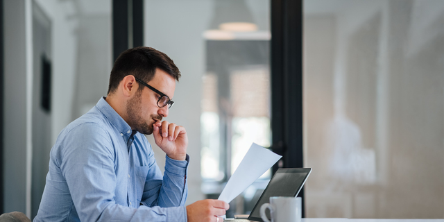 Serious pensive thoughtful young businessman or entrepreneur in modern contemporary office looking at and working with laptop and paper documents making serious and important business decision