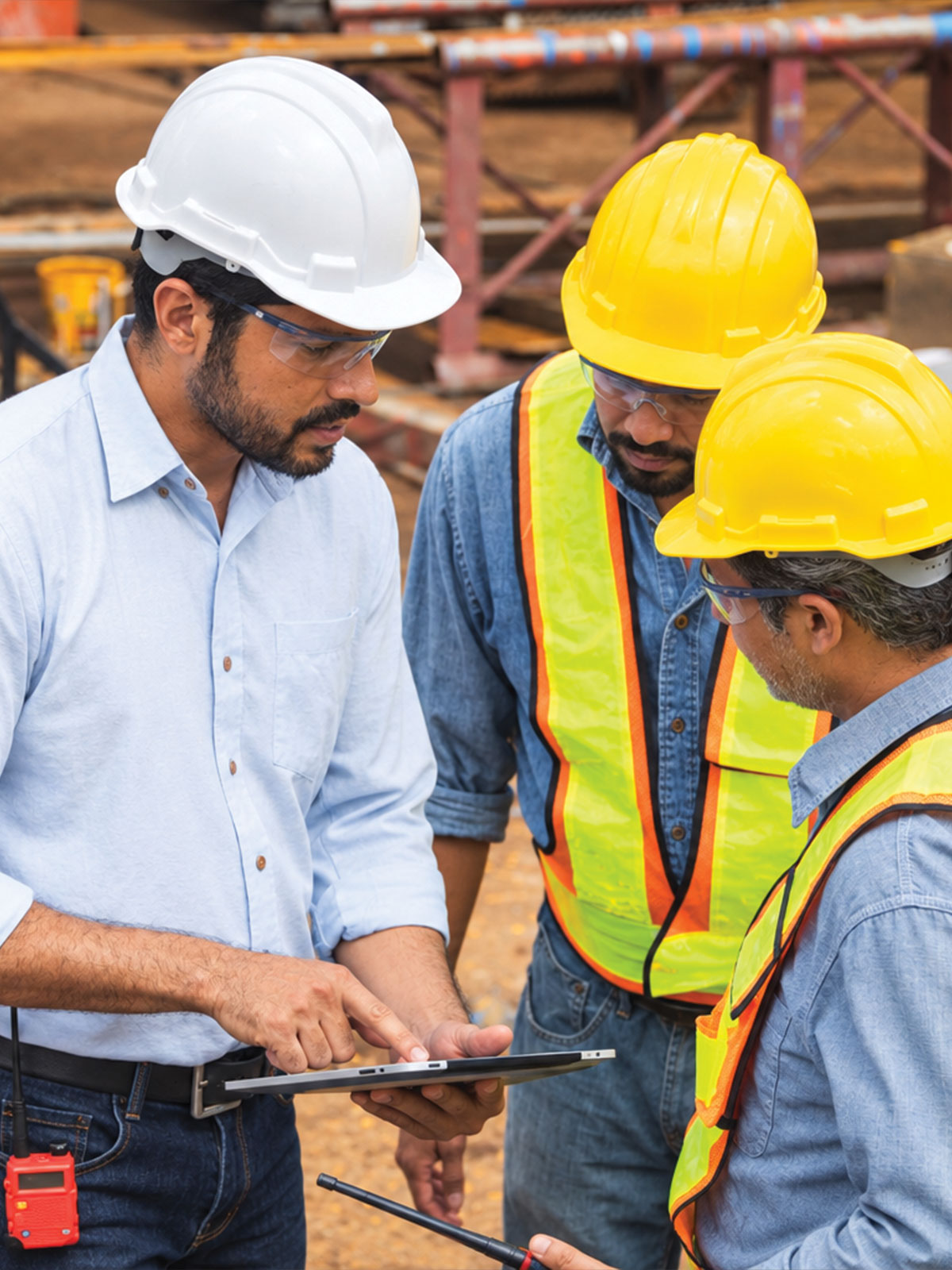 Three telecommunications professionals wearing hard hats, safety glasses, and reflective vests reviewing project plans on a construction site.