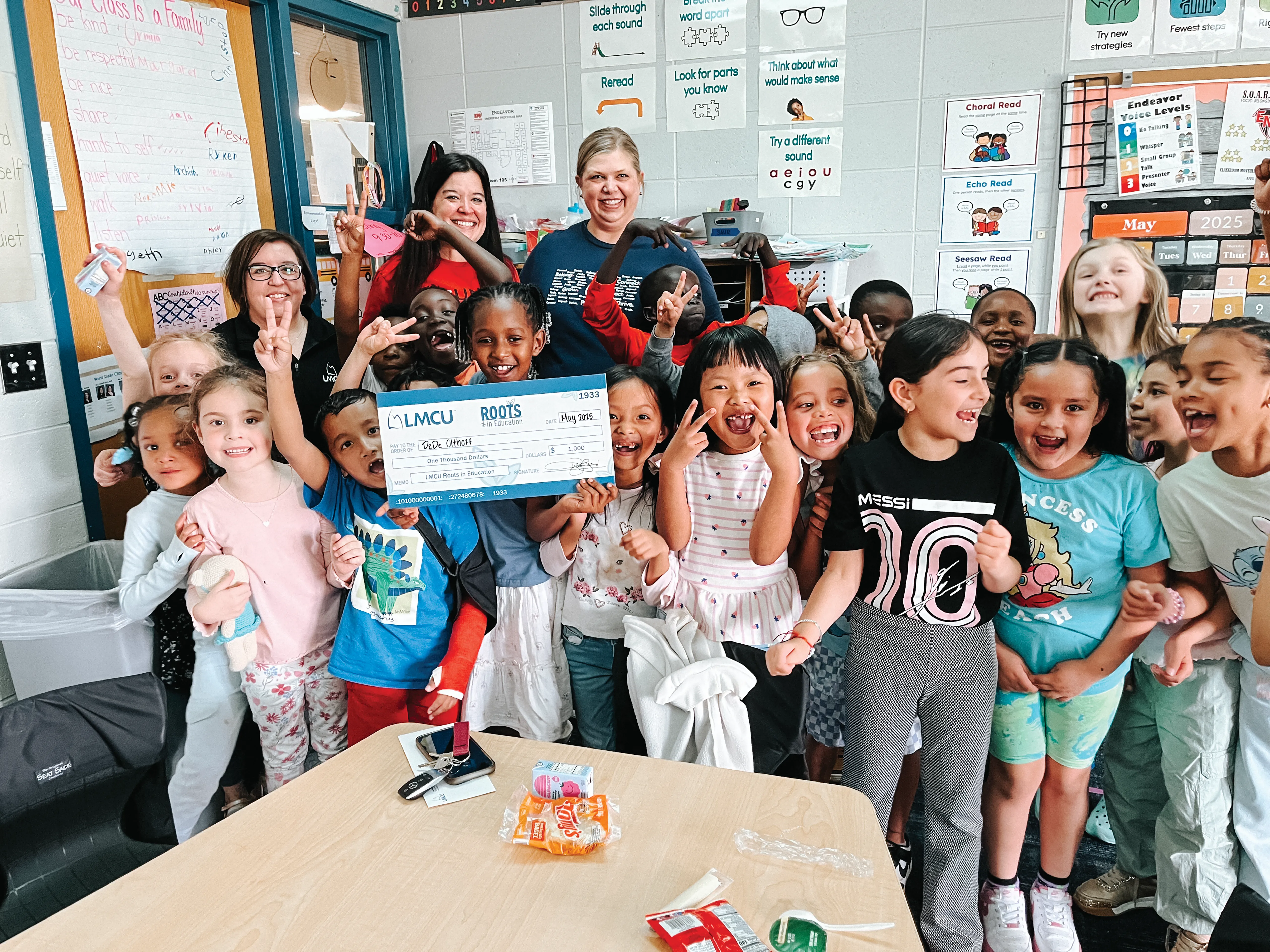 Group of children and two adults posing together in a classroom, with several children holding a large LMCU donation check.