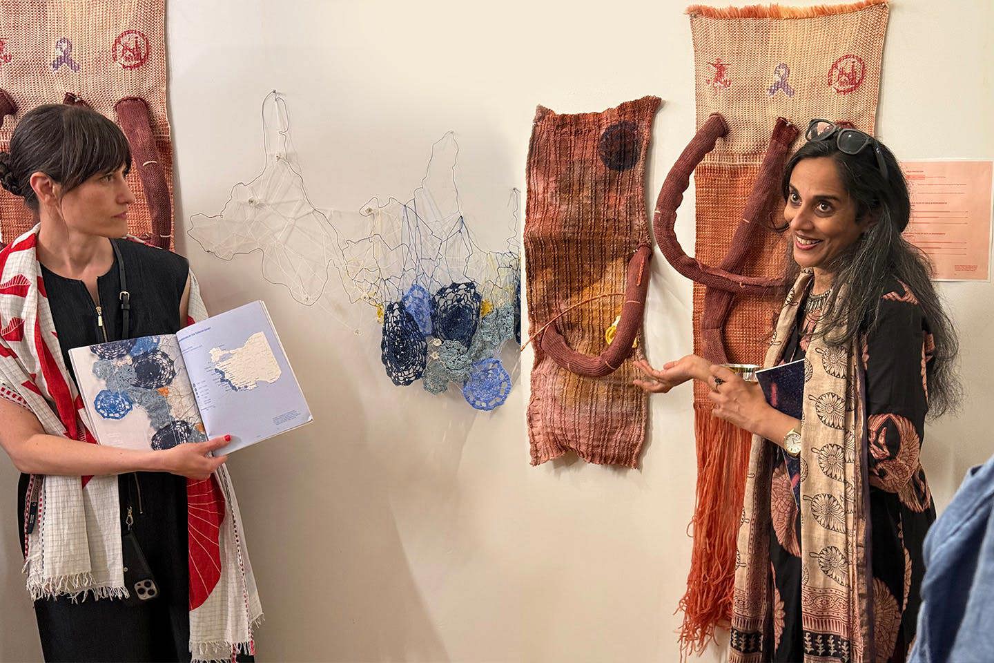 Professors Preeti Gopinath, gesturing toward textiles on the wall, and Anette Millington holding open a book of student work