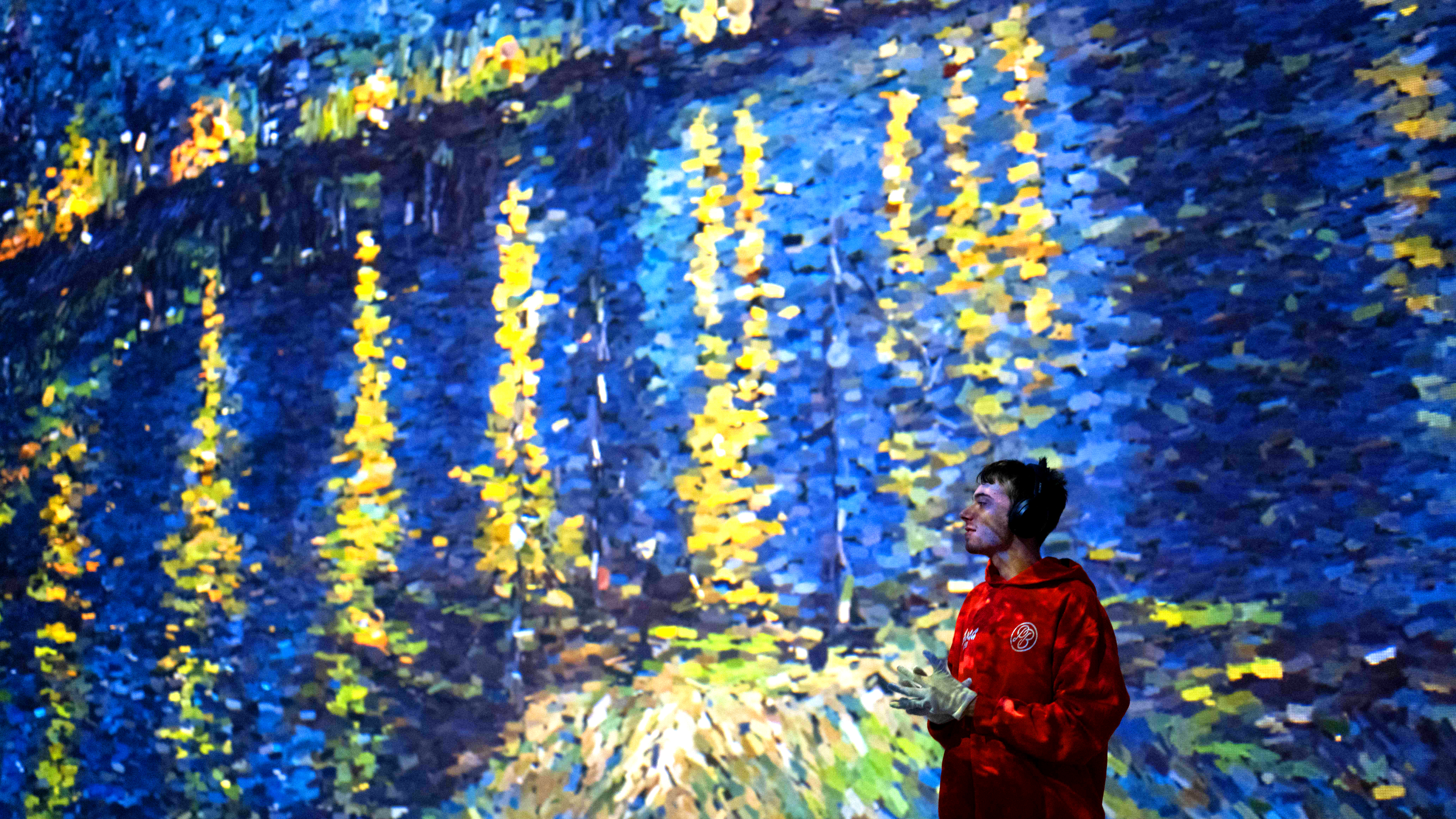 A young man in a red hoodie and gloves stands in front of a large, immersive digital projection of a colourful impressionist-style painting. He wears headphones and smiles while admiring the vibrant blue and yellow display.