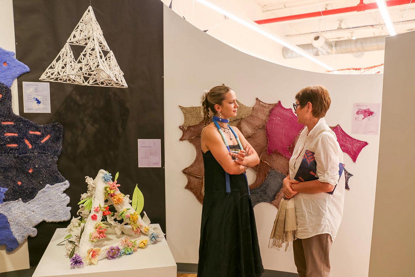 Hattie Batstone stands in front of a display of student projects, engaging in conversation with another viewer at the exhibition.