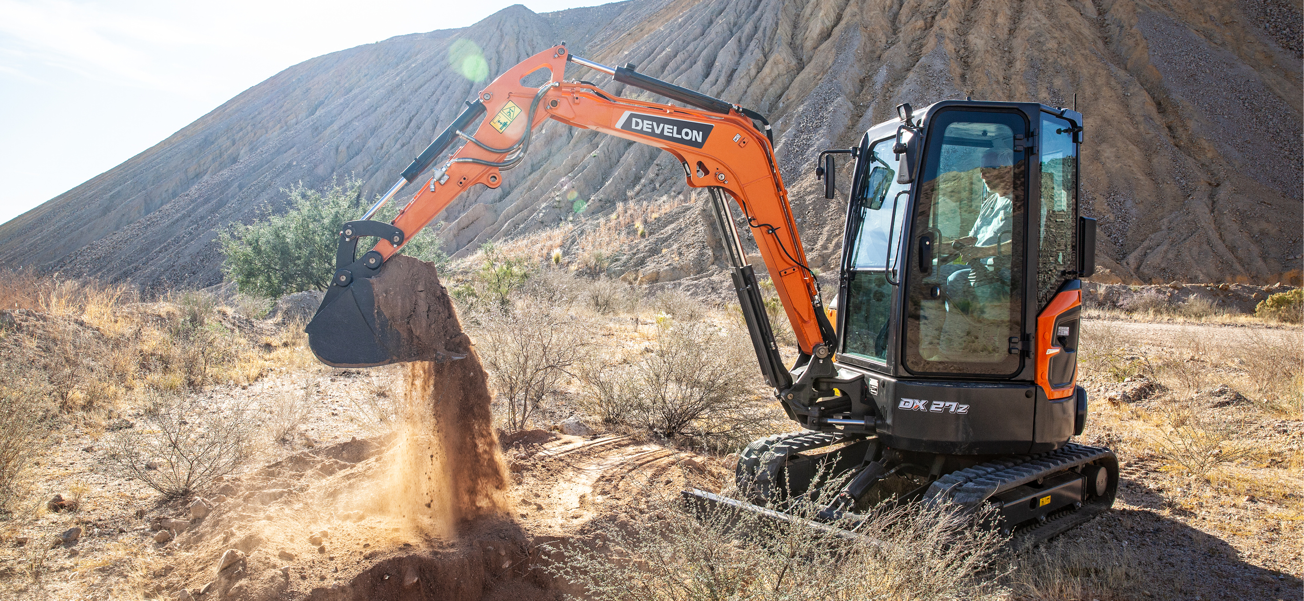 An orange DEVELON DX27Z-7 mini excavator dumping a bucket of dirt in a mountainous landscape.