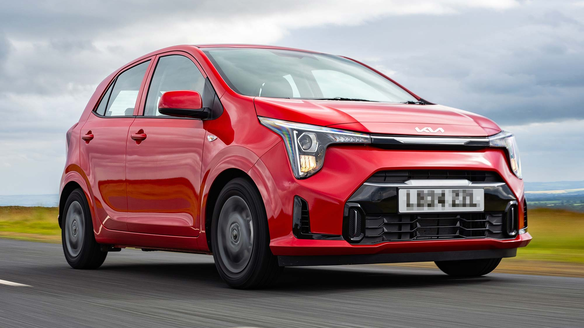 A red Kia Picanto hatchback driving along a countryside road, featuring distinctive headlight design and compact dimensions.