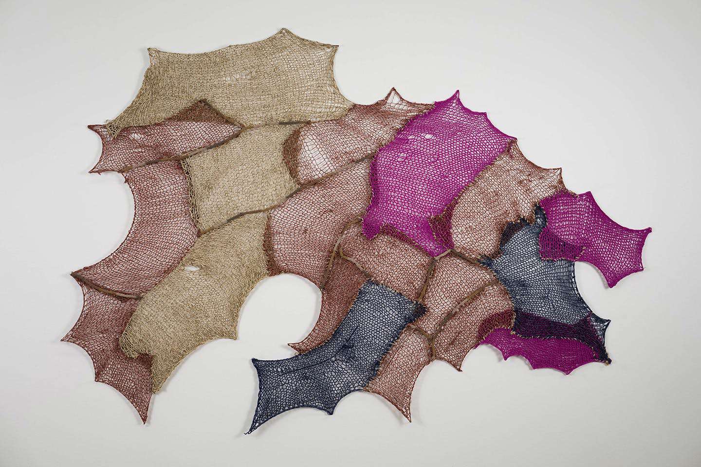 Photograph of an irregularly shaped woven textile artwork by Hattine Batstone, composed of interlocking organic sections in shades of tan, brown, magenta, and navy. The layered, web-like piece feature