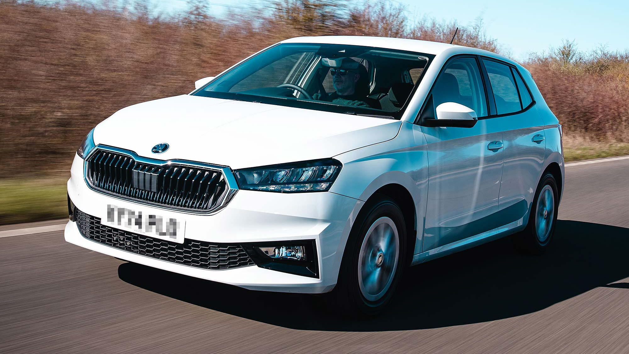 A white Škoda Fabia hatchback driving on an open road, showcasing its sharp body lines and chrome-accented front grille.