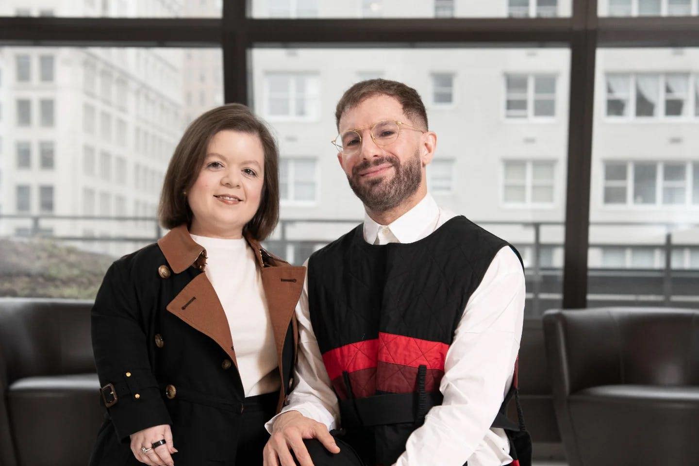 Professor Ben Barry kneels next to disability advocate Sinead Burke in front of windows and furniture in an urban building.
