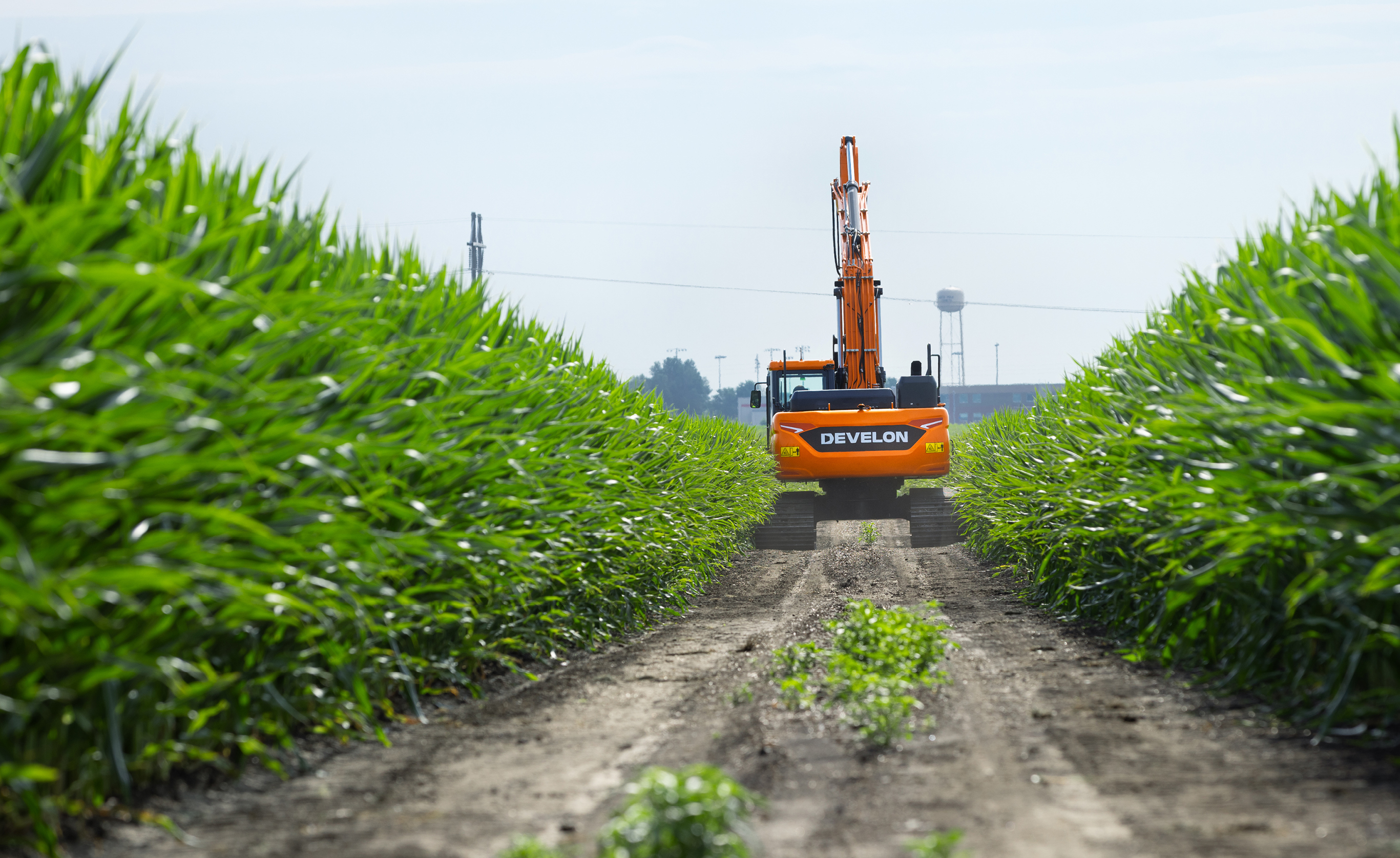 A DEVELON crawler excavator traveling a dirt road surrounded by crops.