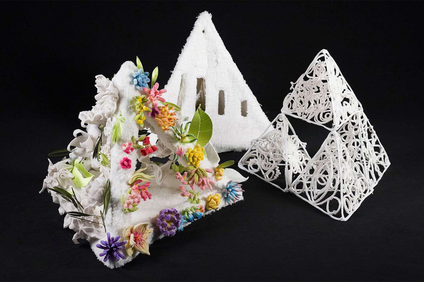 Display photograph of Eva Ziqi Chen’s three textile pyramid structures, adorned with handmade velvet flowers, leaves, and knitted blooms symbolizing the diverse demographics of Thailand’s immigrant po