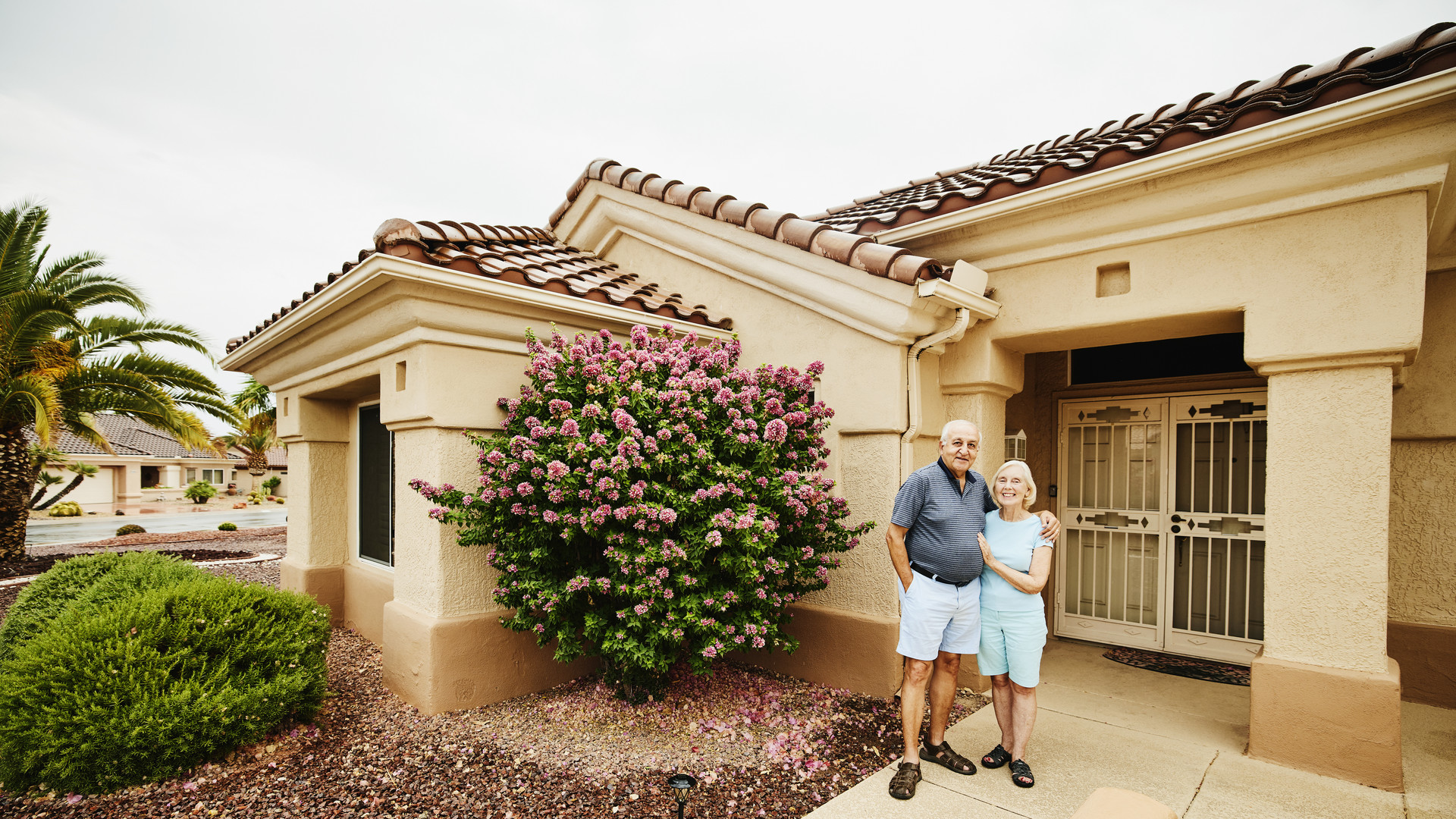 Wide shot of smiling embracing senior couple standing in front of home in retirement community