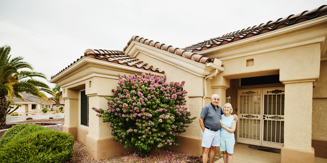 Wide shot of smiling embracing senior couple standing in front of home in retirement community