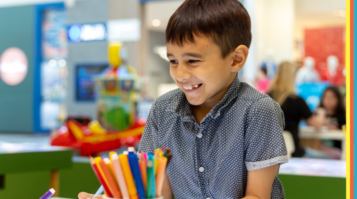 Smiling young child drawing with coloured pencils at a table in a shopping centre play area, with a blurred family-friendly background and colourful pencils in the foreground