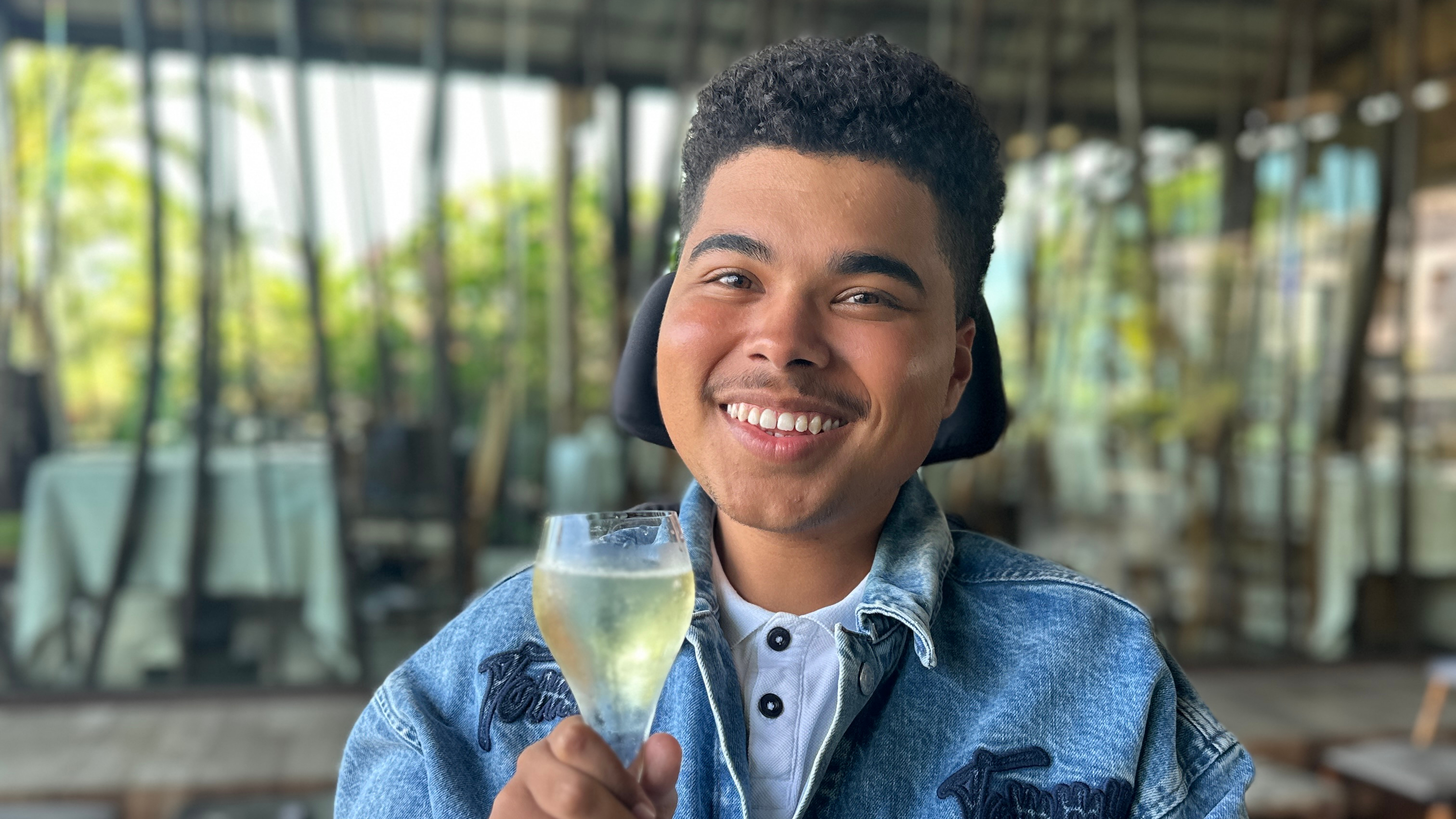 Maxwell McKnight smiles and raises a glass at a restaurant while seated in his powered wheelchair, wearing a denim jacket, with soft lighting and a modern interior in the background.