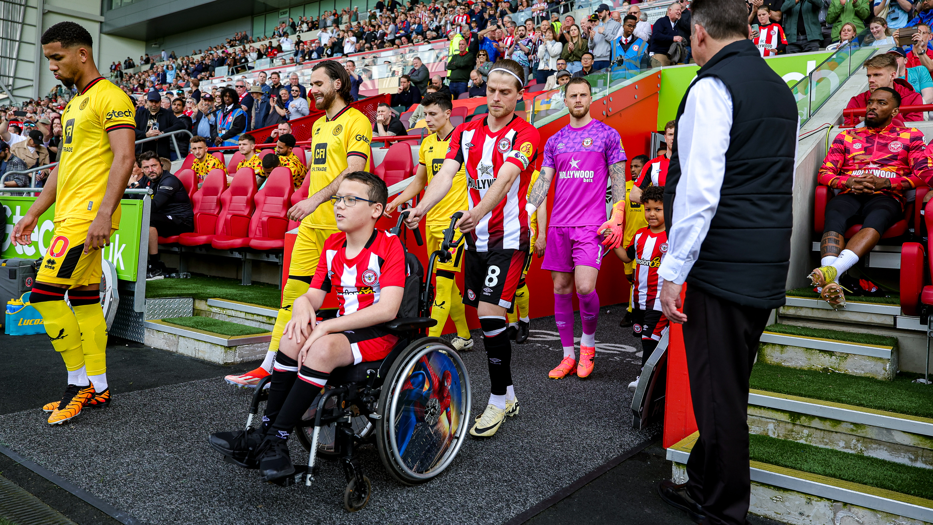 A young Brentford FC fan in a wheelchair leads the football team onto the pitch alongside players in red-and-white and yellow kits. The stadium is filled with spectators, and team members walk out from the dugout behind him.