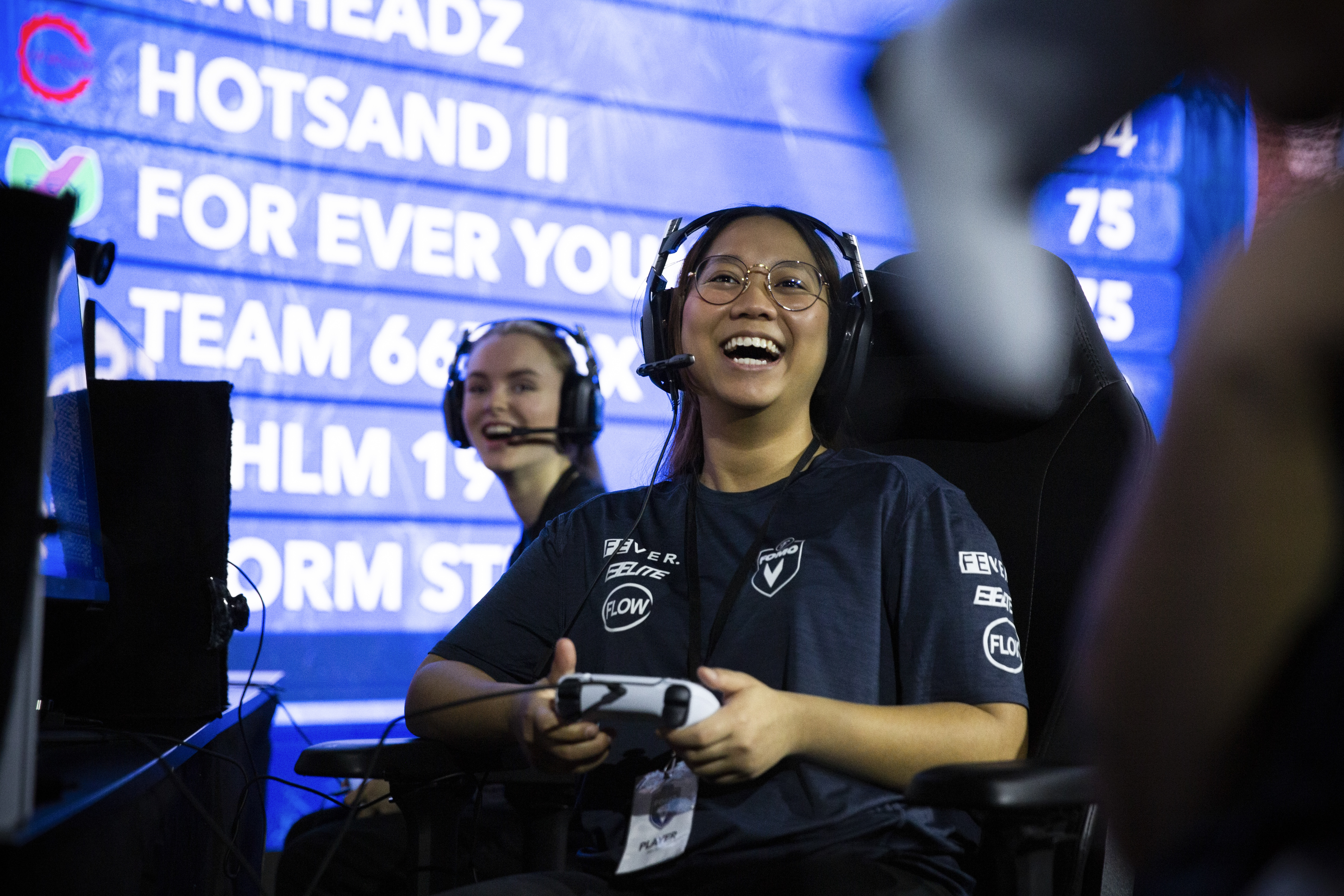 A player in an esports venue smiles with a controller in hand, wearing a headset in front of a scoreboard.