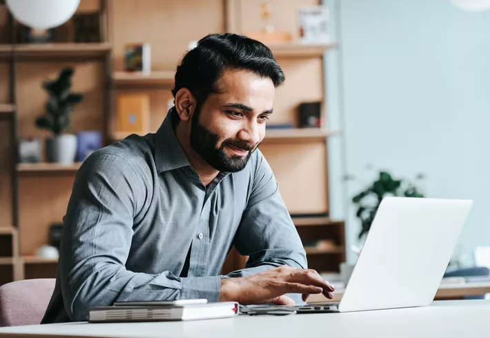 Smiling Middle Eastern professional seated at desk looking at laptop 