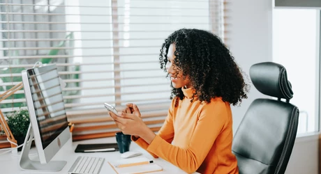 Professional African American woman sitting at desk in office looking at cell phone 