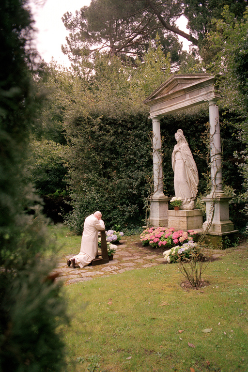 John Paul II kneeling with head down in prayer in front of a statue of our lady outside in a garden