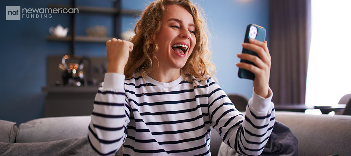 young, curly haired woman celebrating while looking a mobile phone