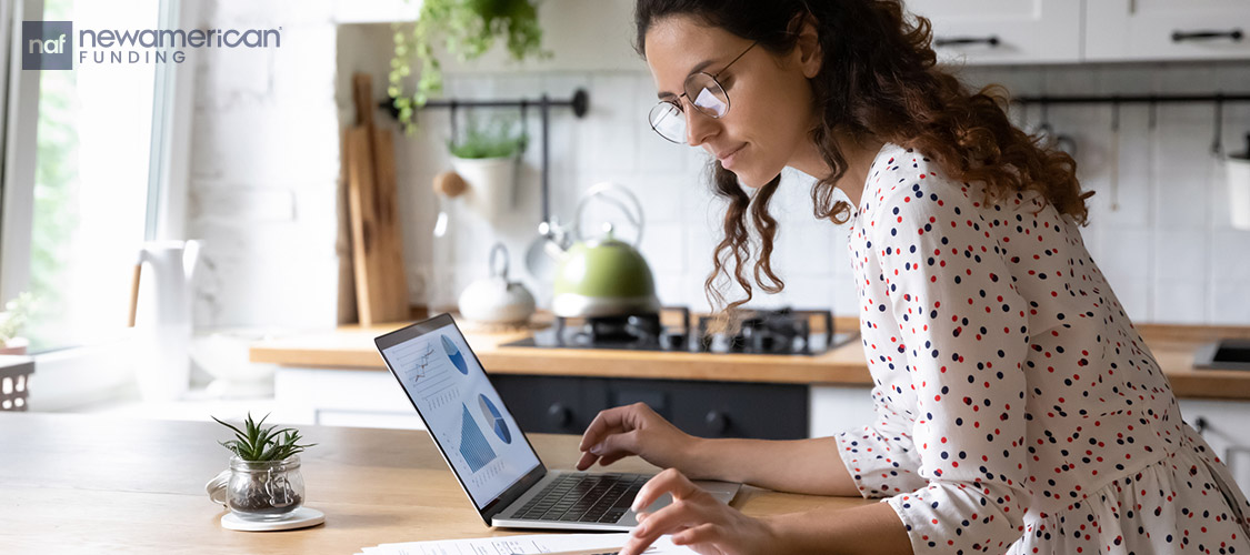 woman using a calculator while on a laptop in the kitchen