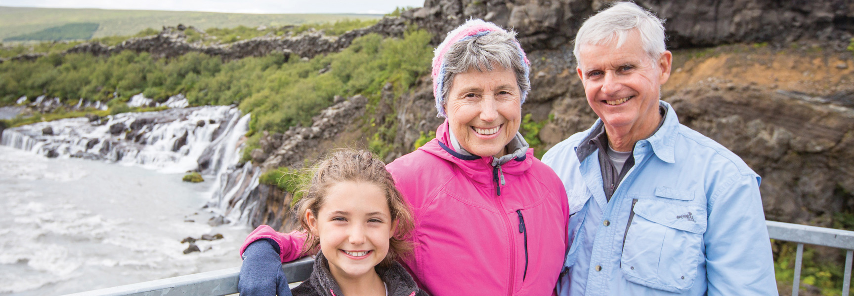 A smiling girl with her grandparents stand in front of a waterfall in Iceland.